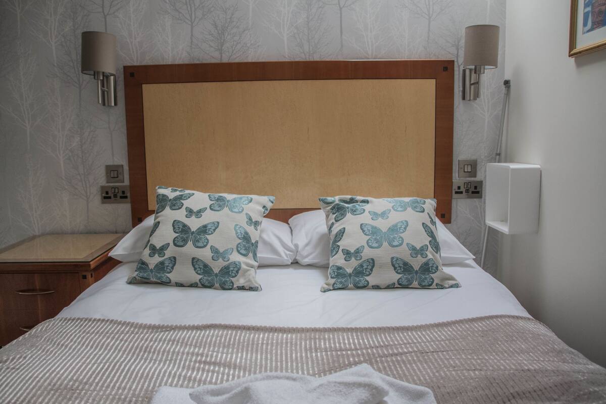 A hotel room with a wooden headboard, two butterfly-patterned pillows, a white bedspread, and a bedside table with a lamp.