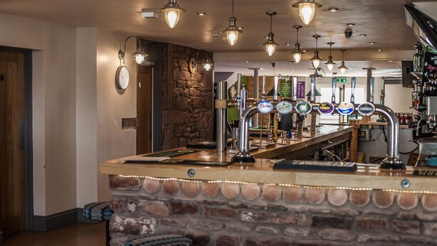 A bar area with a stone counter, multiple taps, and hanging light fixtures. - The Lion and The Lamb (Seascale)