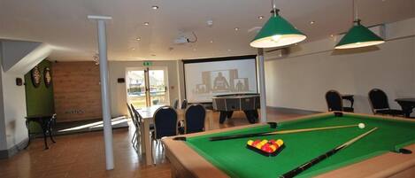A pool table with balls, a projector screen, and a billiards table in a room with chairs and a table.