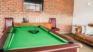 A pool table in a room with a brick wall, a television mounted above it, and a leather sofa with cushions. - Keld Green House (Seascale)