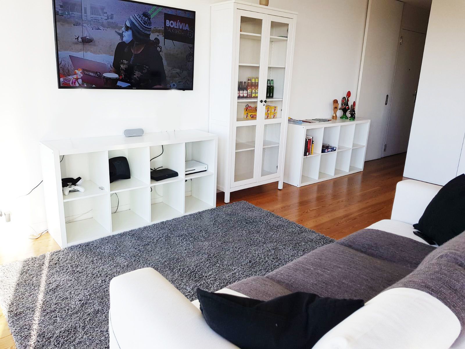 A modern living room with a flat-screen TV, white shelving units, and a grey carpet.