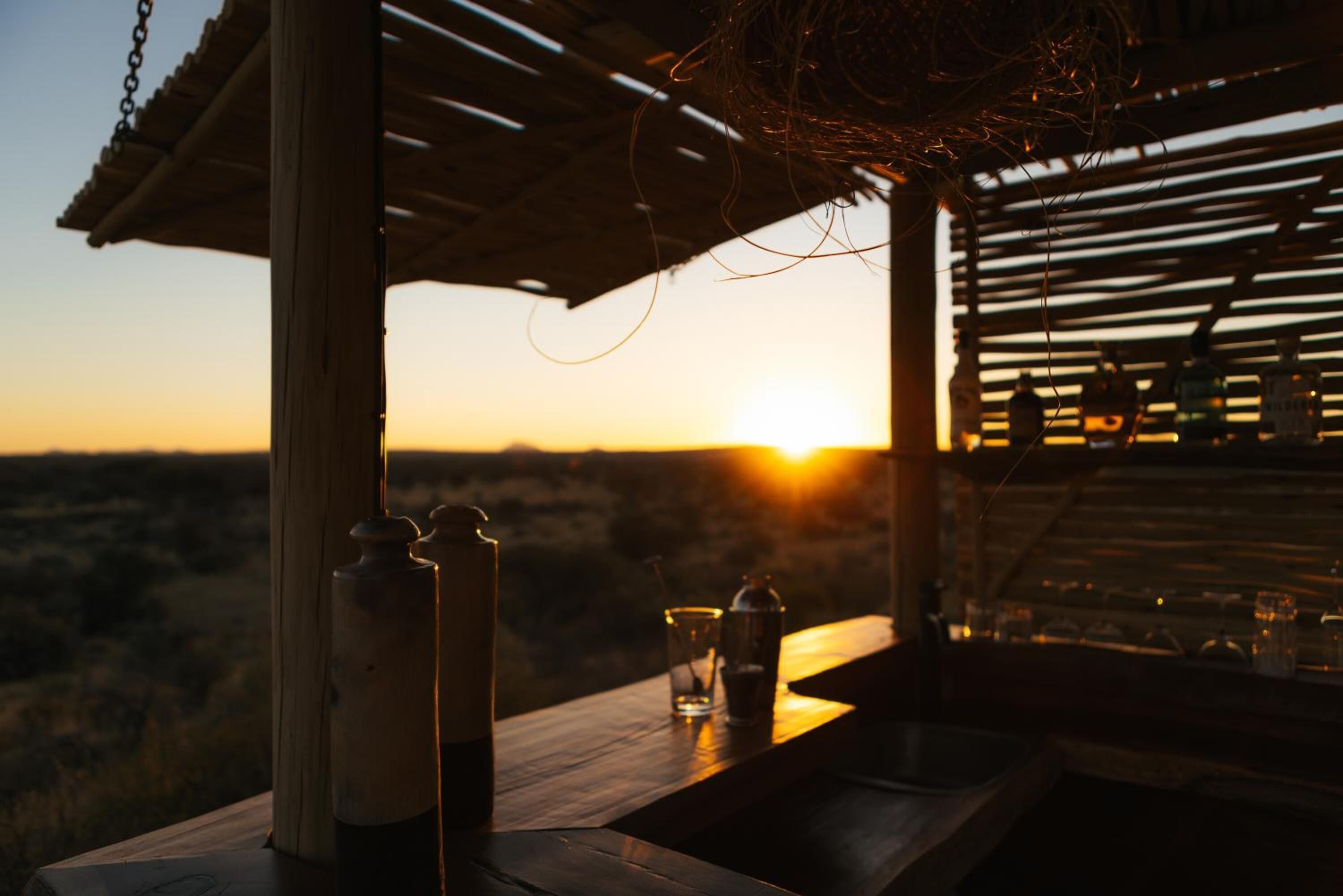 A wooden outdoor seating area with a view of the sunset, a bottle, and a glass on the table.