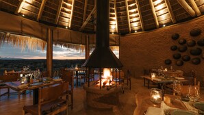 A restaurant with a thatched roof, wooden furniture, and a central wood-burning stove. Tables are set for dining with wine glasses and cutlery. -  Zannier Omaanda (Windhoek)