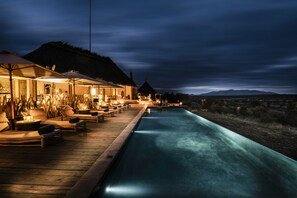 A poolside area with lounge chairs and umbrellas, a thatched-roof building, and a view of distant mountains. -  Zannier Omaanda (Windhoek)