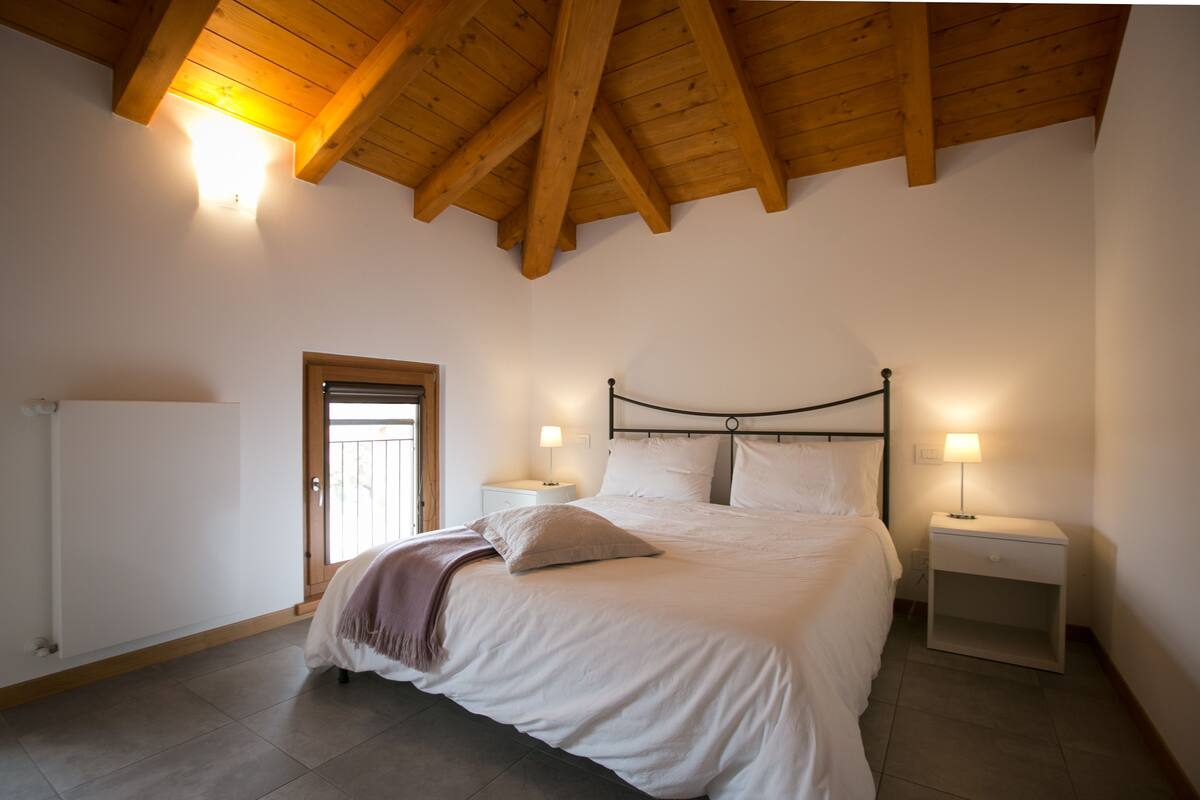 Quarto com teto de madeira, cama com roupa de cama branca, duas mesas de cabeceira com abajures e uma porta à esquerda.