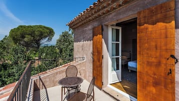 A balcony with a table and chairs, a wooden door, and a view of a tree and greenery.