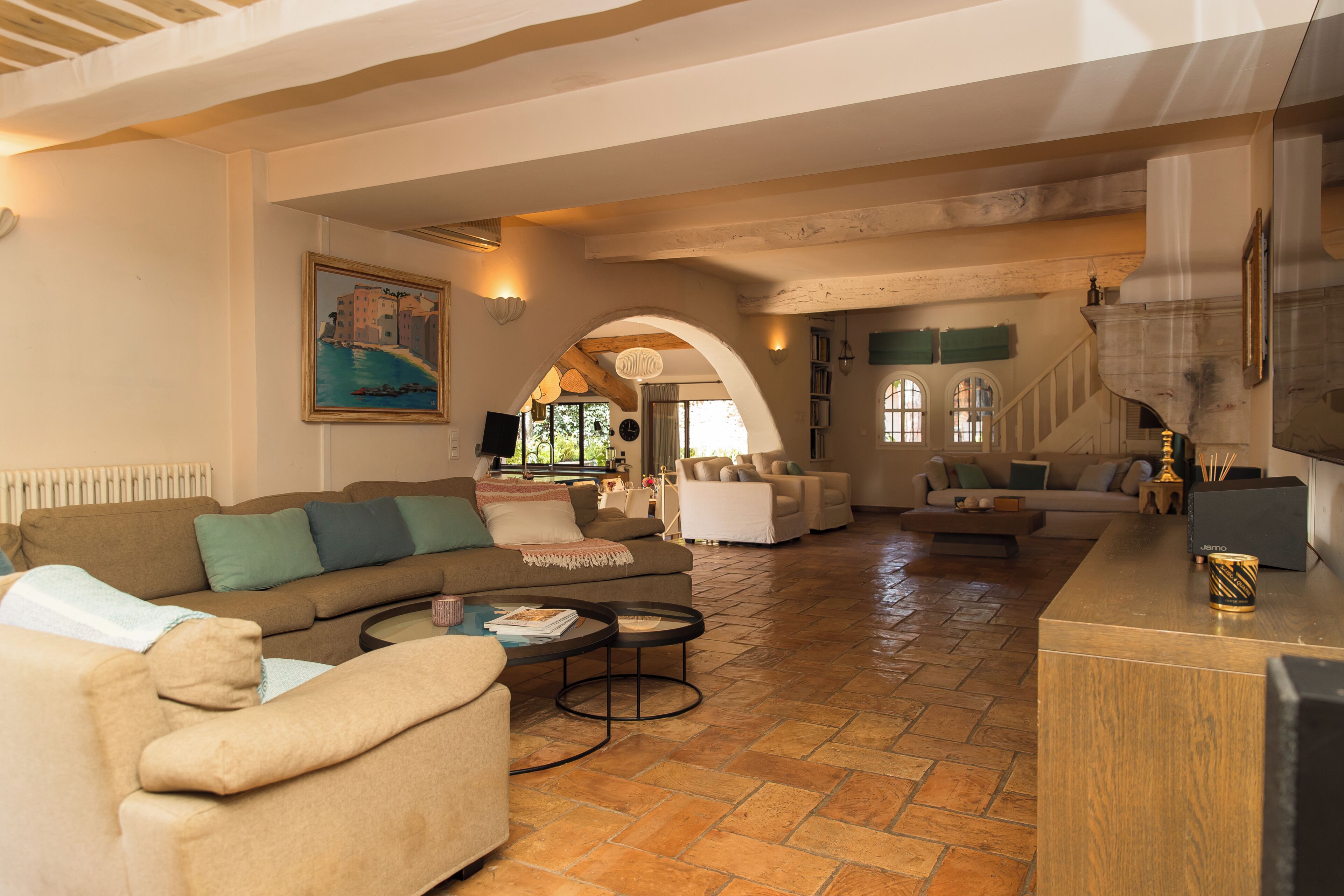 A spacious living area with a large sofa, armchairs, and a coffee table. There is a fireplace, a painting on the wall, and a staircase leading to another level.