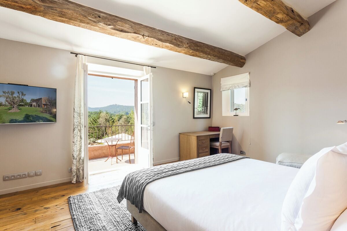 A bedroom with a large bed, a desk, a chair, a TV, and a balcony with a view of the outdoors.