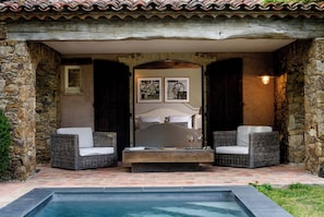A stone-walled bedroom with a bed, two wicker sofas, a small table, and a swimming pool. - Domaine Les Mésanges (Gassin)