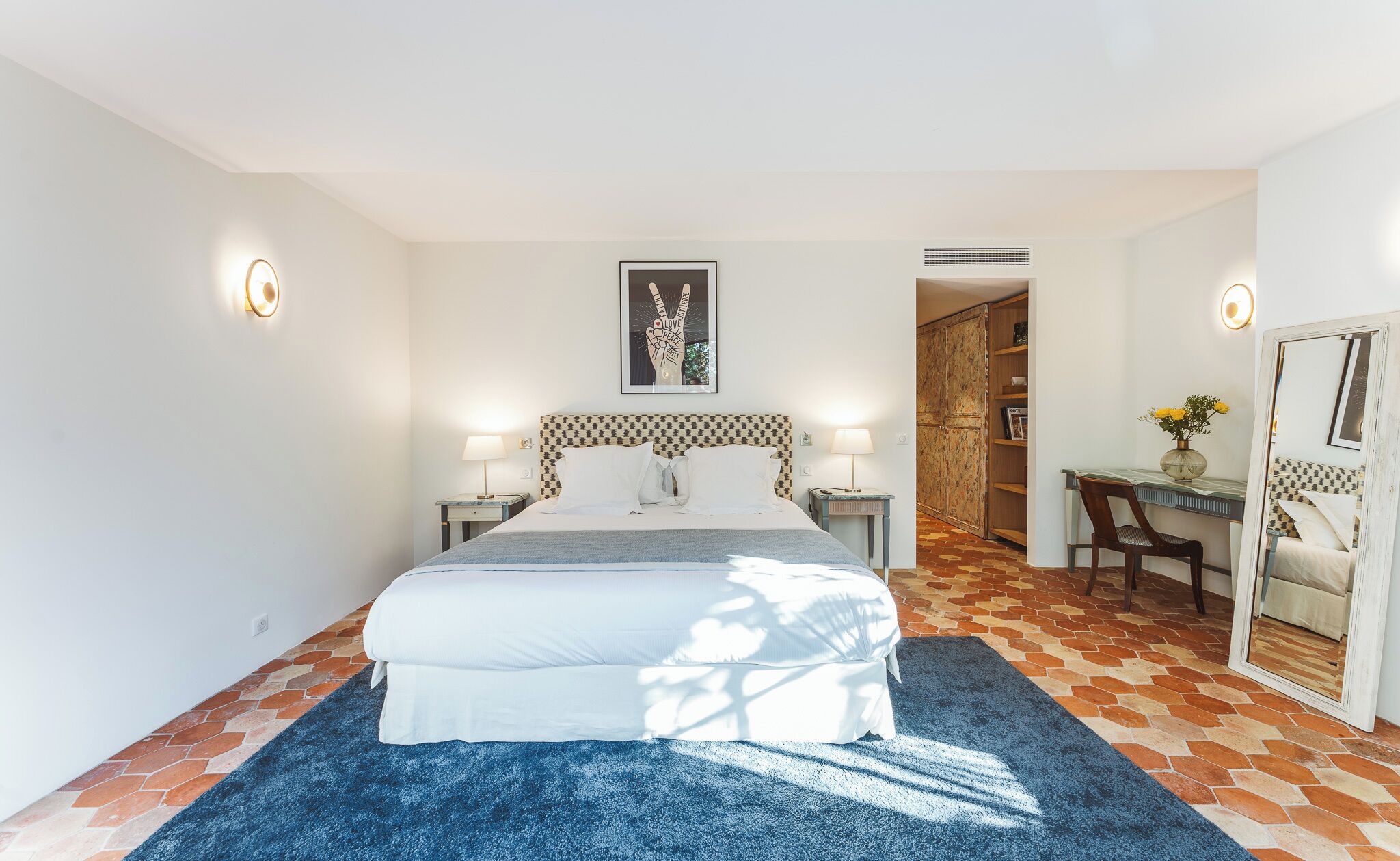A bedroom with a large bed, bedside tables with lamps, a desk with a chair, a wardrobe, and a framed picture on the wall.
