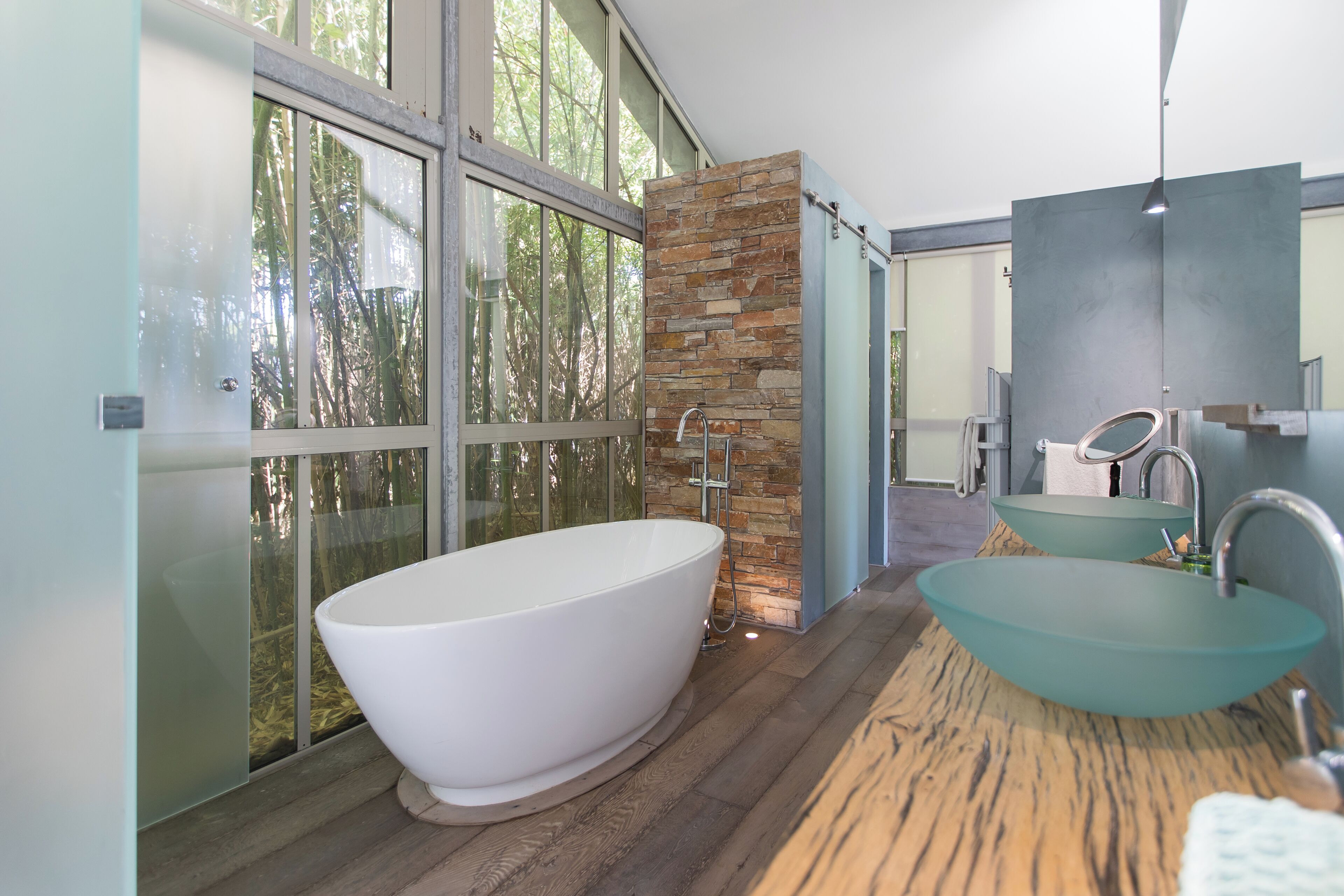 A modern bathroom with a freestanding bathtub, a stone accent wall, and a large window offering a view of trees.