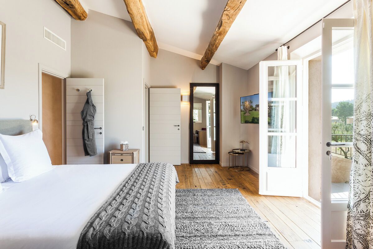 A bedroom with a large bed, wooden beams, a door leading to a balcony, and a view of the outdoors.