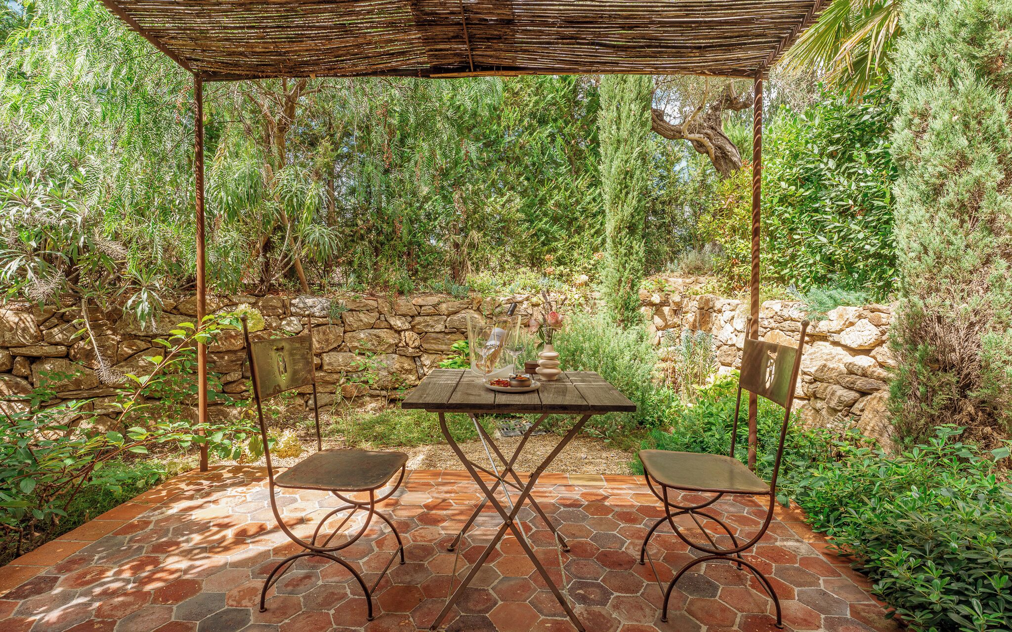 A covered patio with a table and two chairs, surrounded by greenery and stone walls.