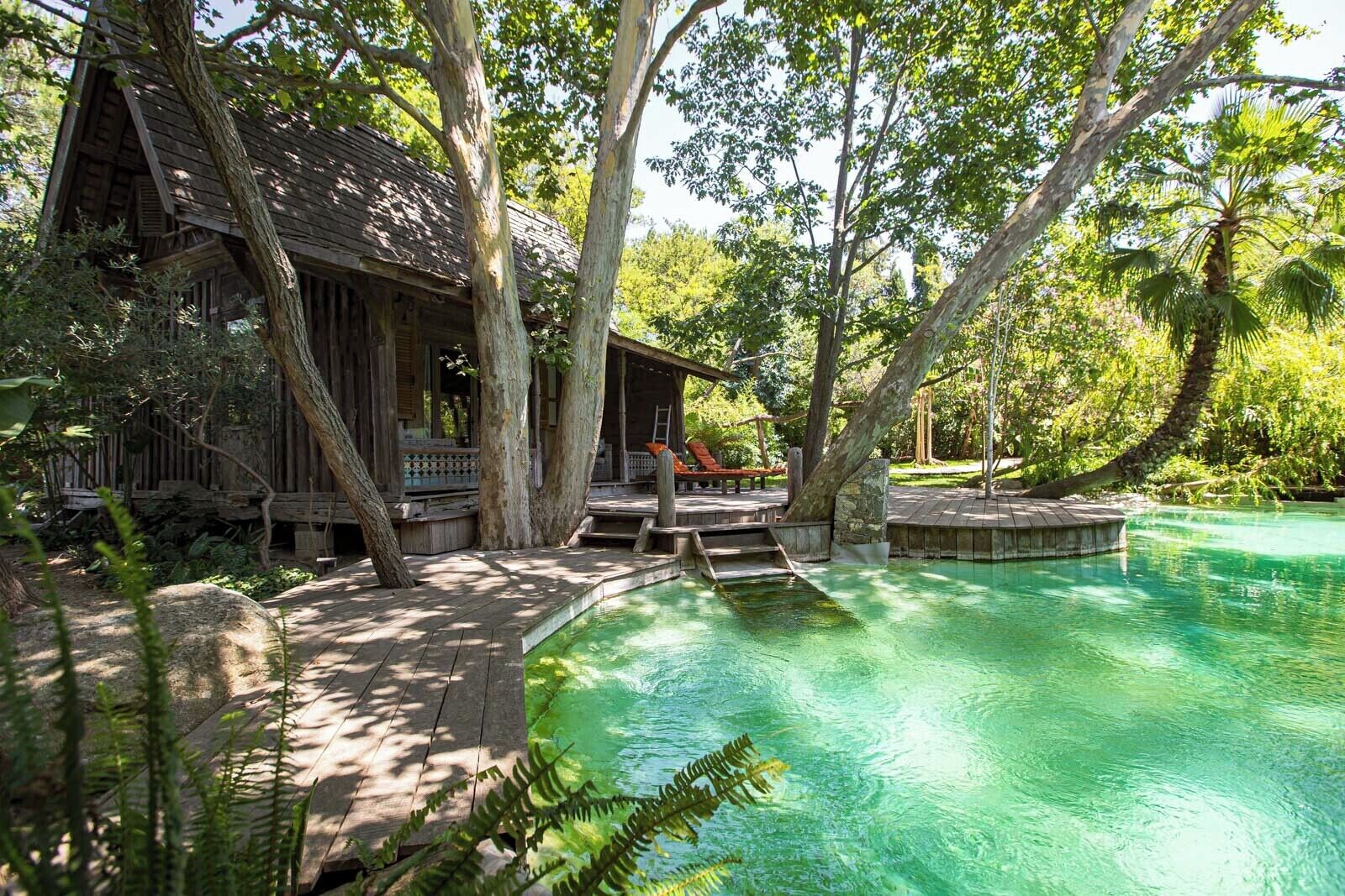 A wooden cabin with a deck, surrounded by trees and a clear pool.