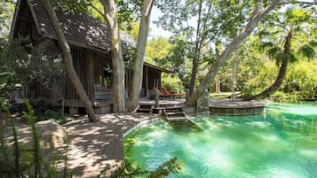 A wooden cabin with a deck, surrounded by trees and a clear pool.