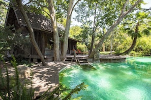 Una cabaña de madera con terraza, rodeada de árboles y una piscina cristalina.