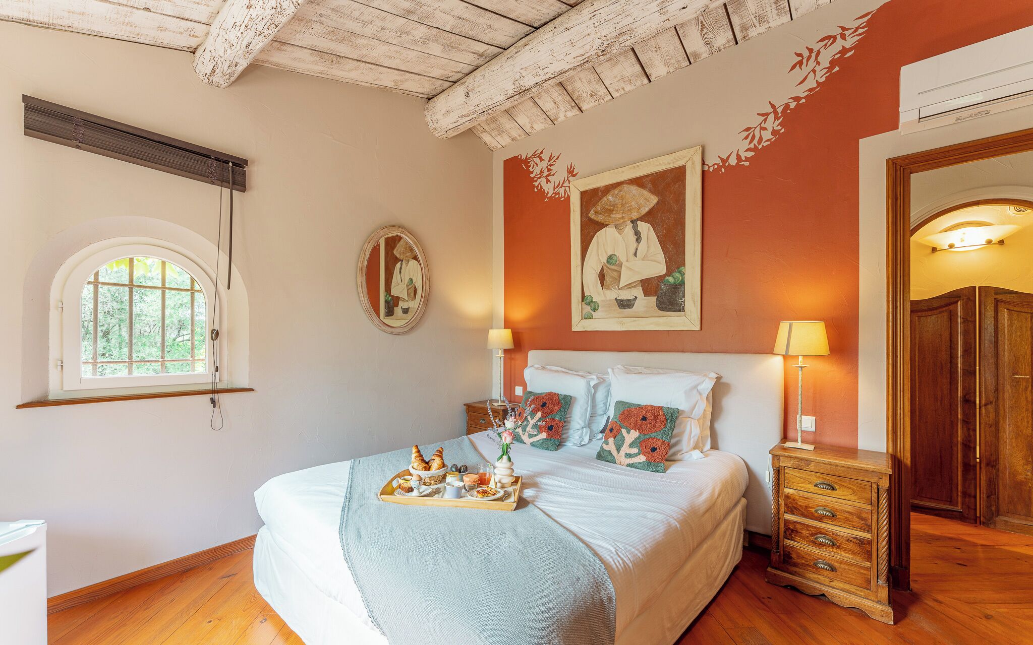 A bedroom with a large bed, bedside lamps, a wooden nightstand, a framed picture on the wall, and a window with a view of greenery.