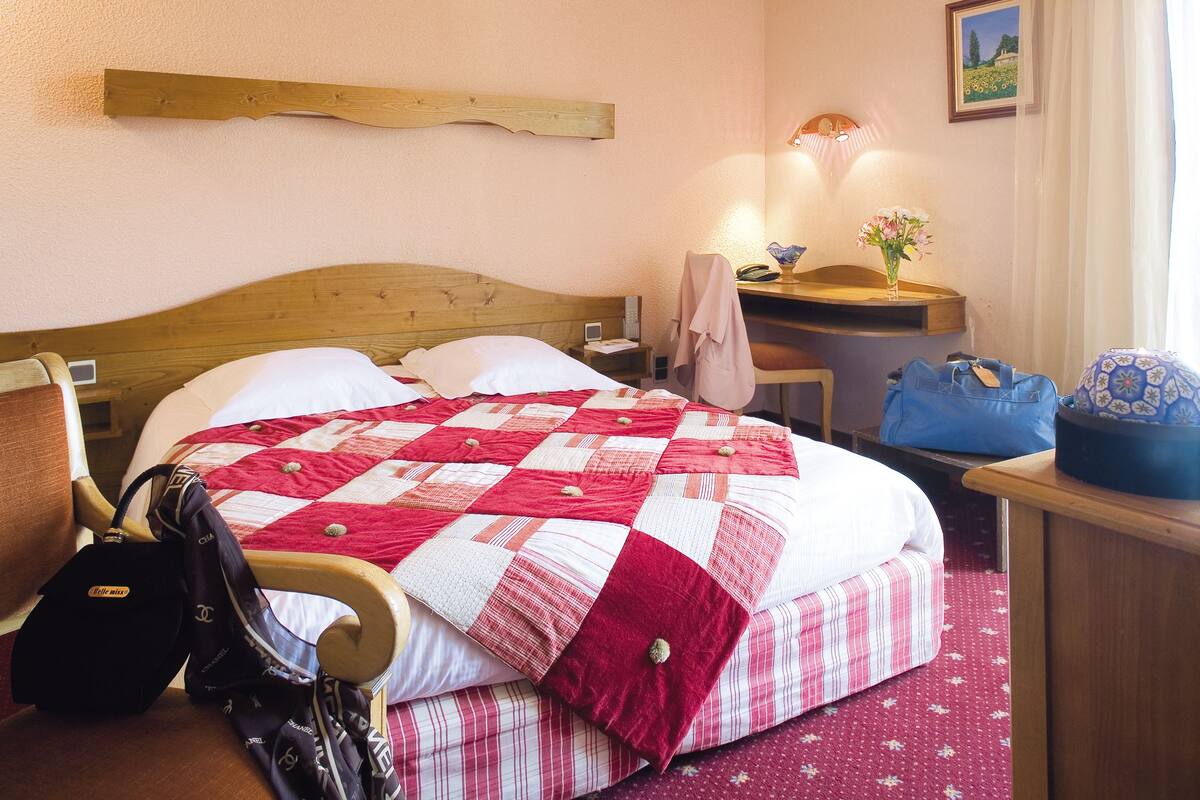 Hotellihuone, jossa on puinen sänky, kirjoituspöytä lampun kanssa, tuoli ja matkalaukku.