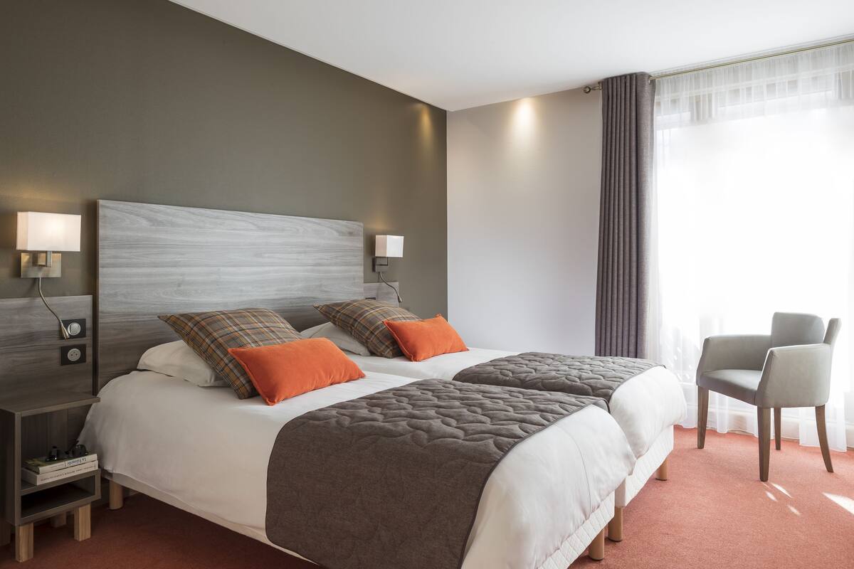 Hotellihuone, jossa on kaksi sänkyä, yöpöytä, tuoli ja suuri ikkuna, jossa on verhot.
