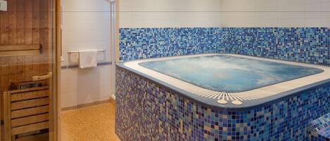 Un'area benessere con vasca idromassaggio rivestita in mosaico, una porta in legno e pareti piastrellate di bianco.