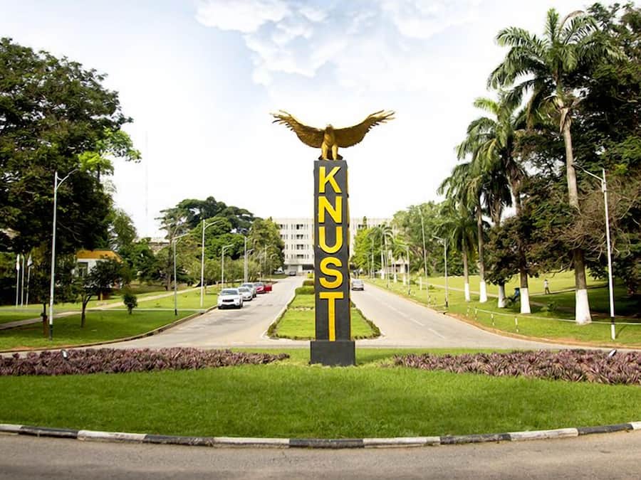 Un panneau de signalisation orné d’une statue d’aigle et du mot « KNUST », le tout entouré de verdure et de palmiers.