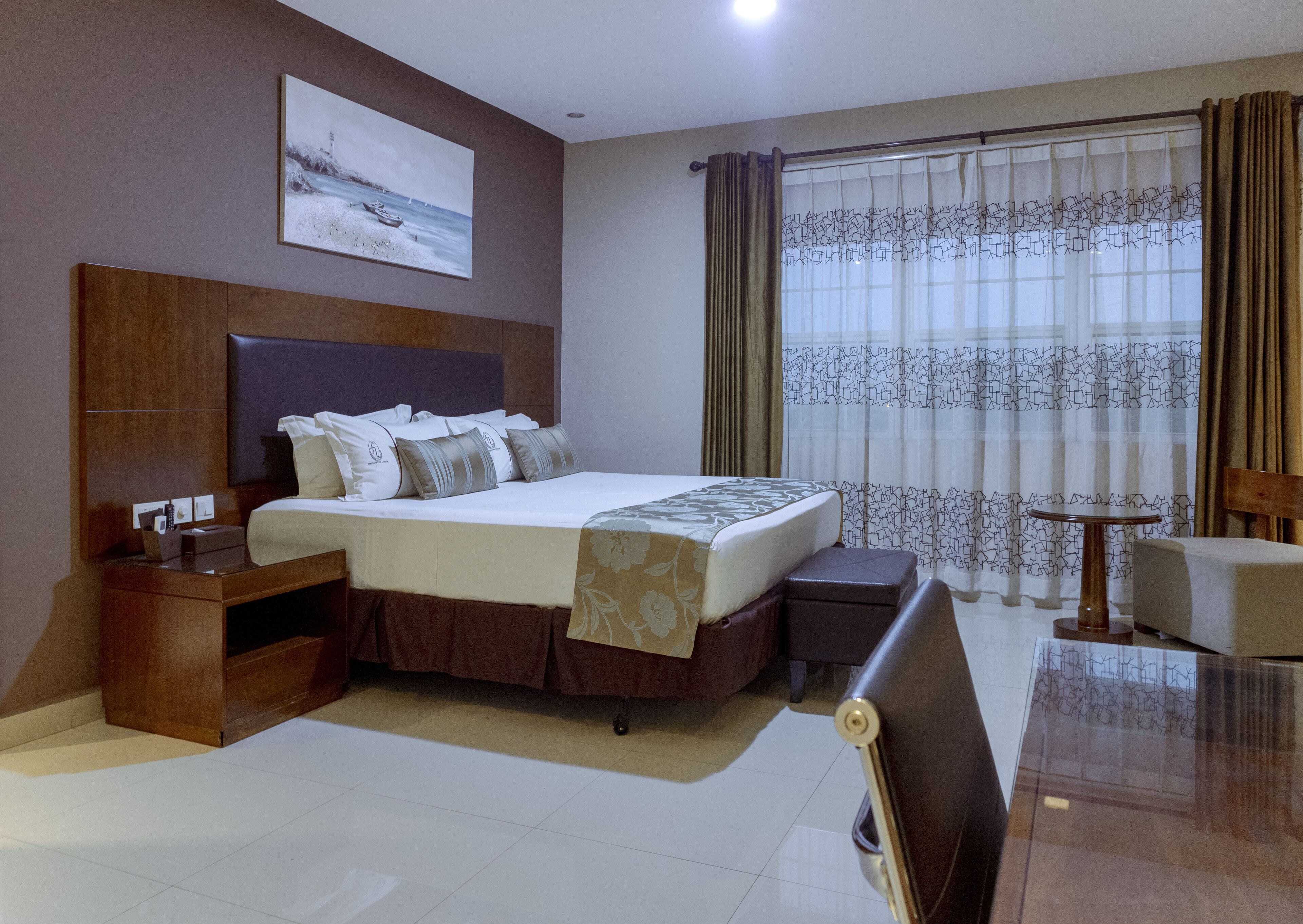 A hotel room with a large bed, a wooden headboard, a bedside table with a lamp, a small table, and a chair.