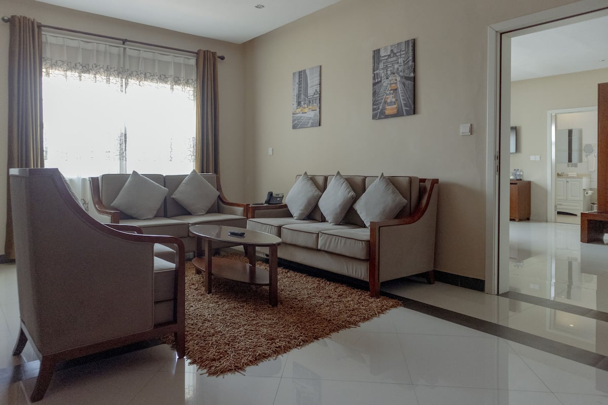 A modern living room with a sofa, armchair, and coffee table. There are two paintings on the wall and a window with curtains.