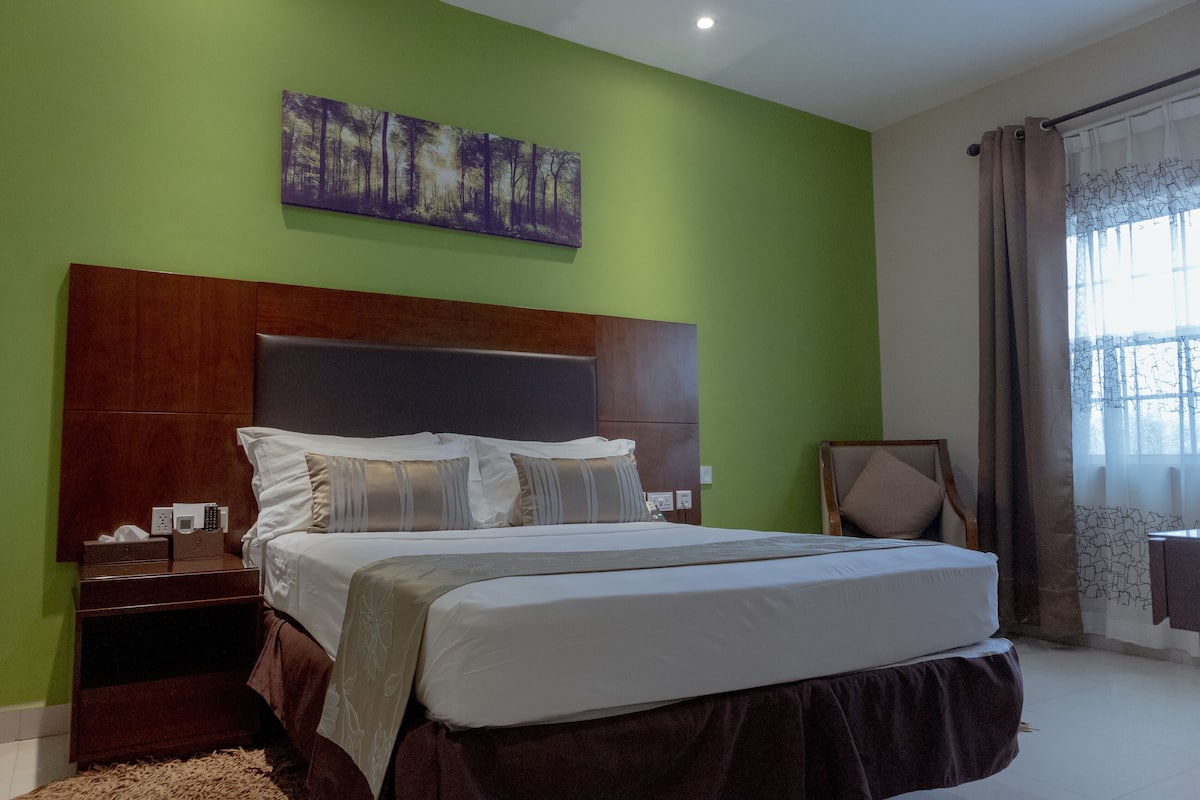 A bedroom with a large bed, a nightstand, a wall-mounted painting, and a window with curtains.