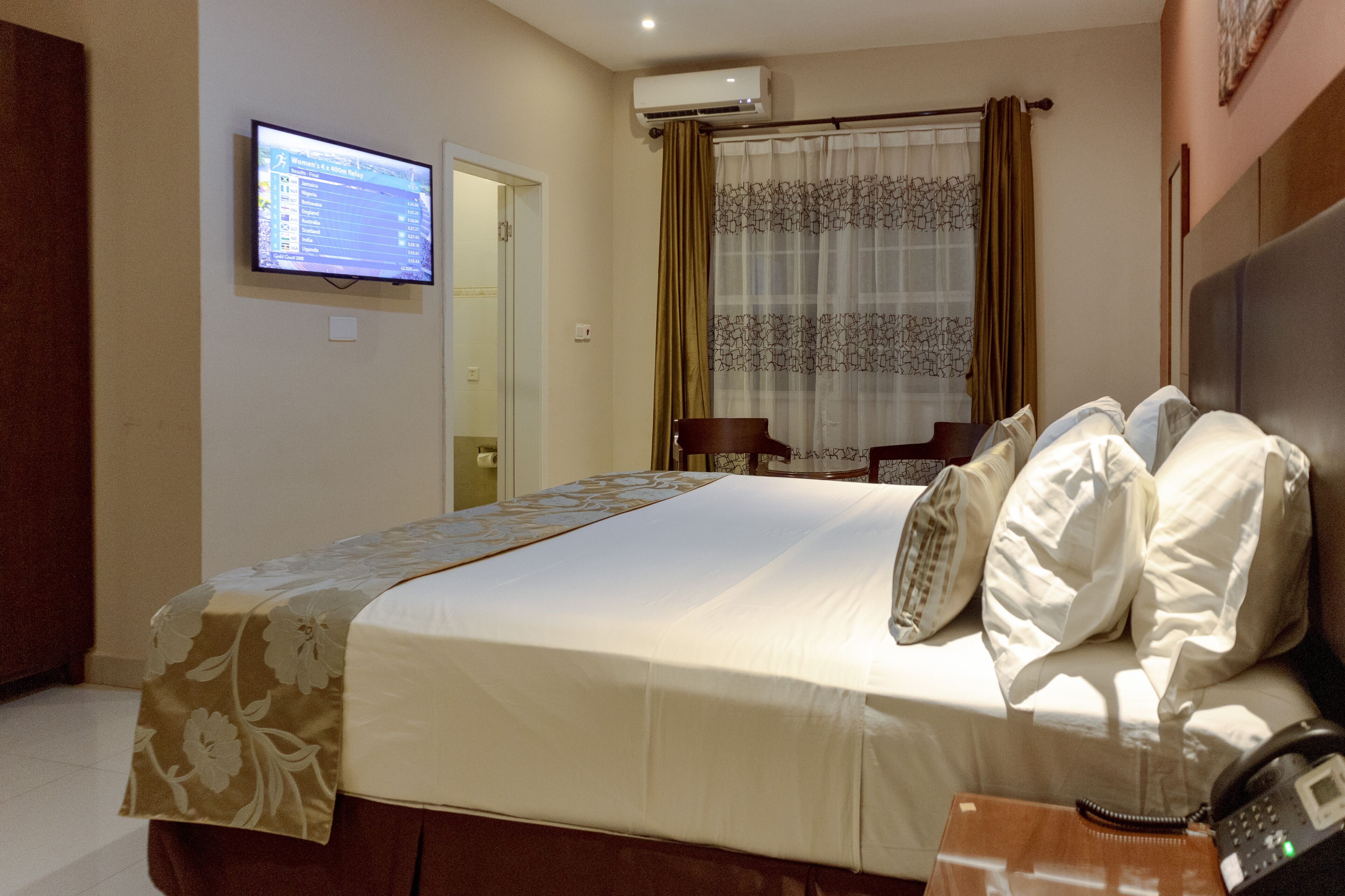 Ett hotellrum med en säng, en telefon vid sängen, en tv som är monterad på väggen och ett fönster med gardiner.