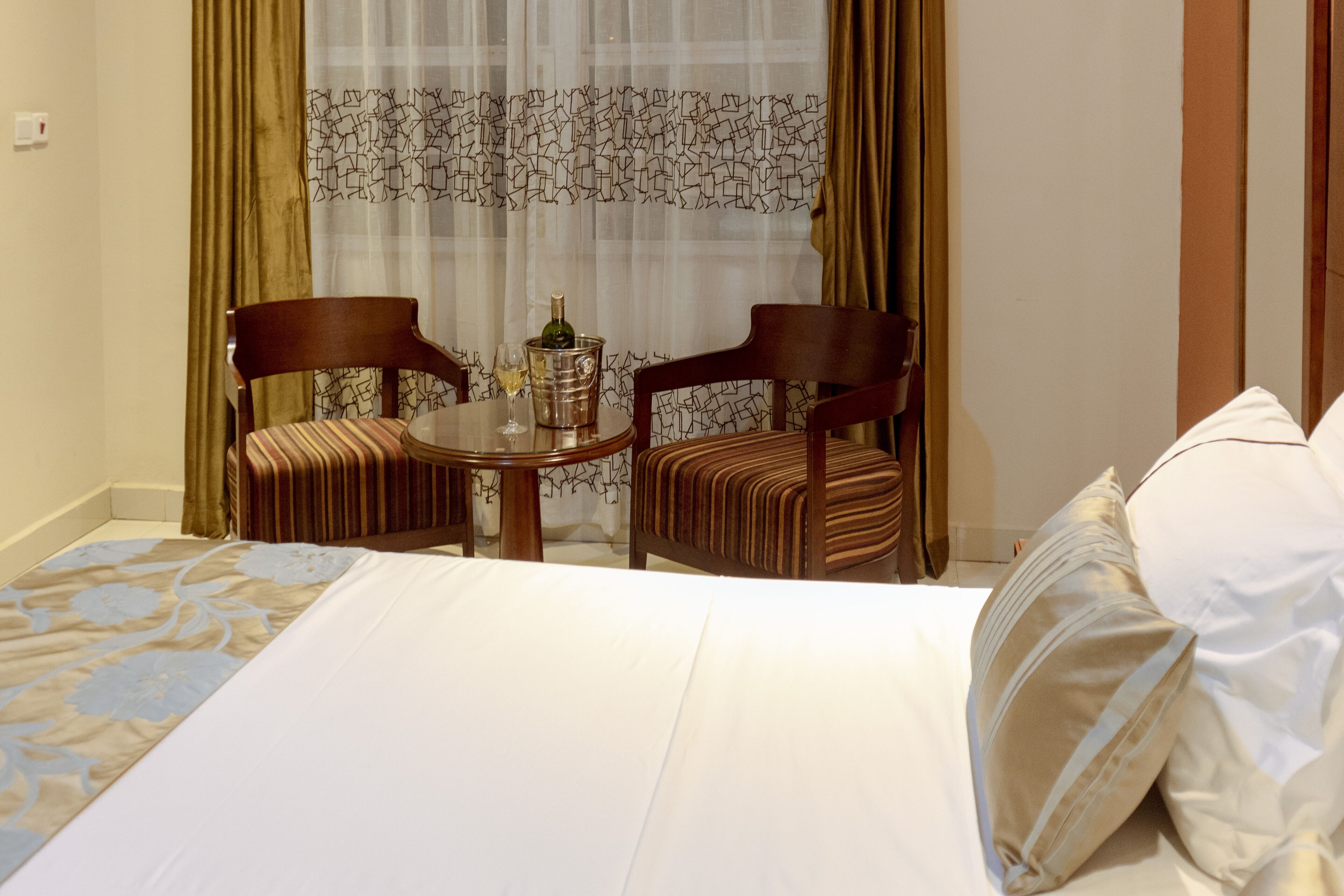 A hotel room with a bed, two upholstered chairs, a small round table with a bottle and glass, and a patterned curtain.