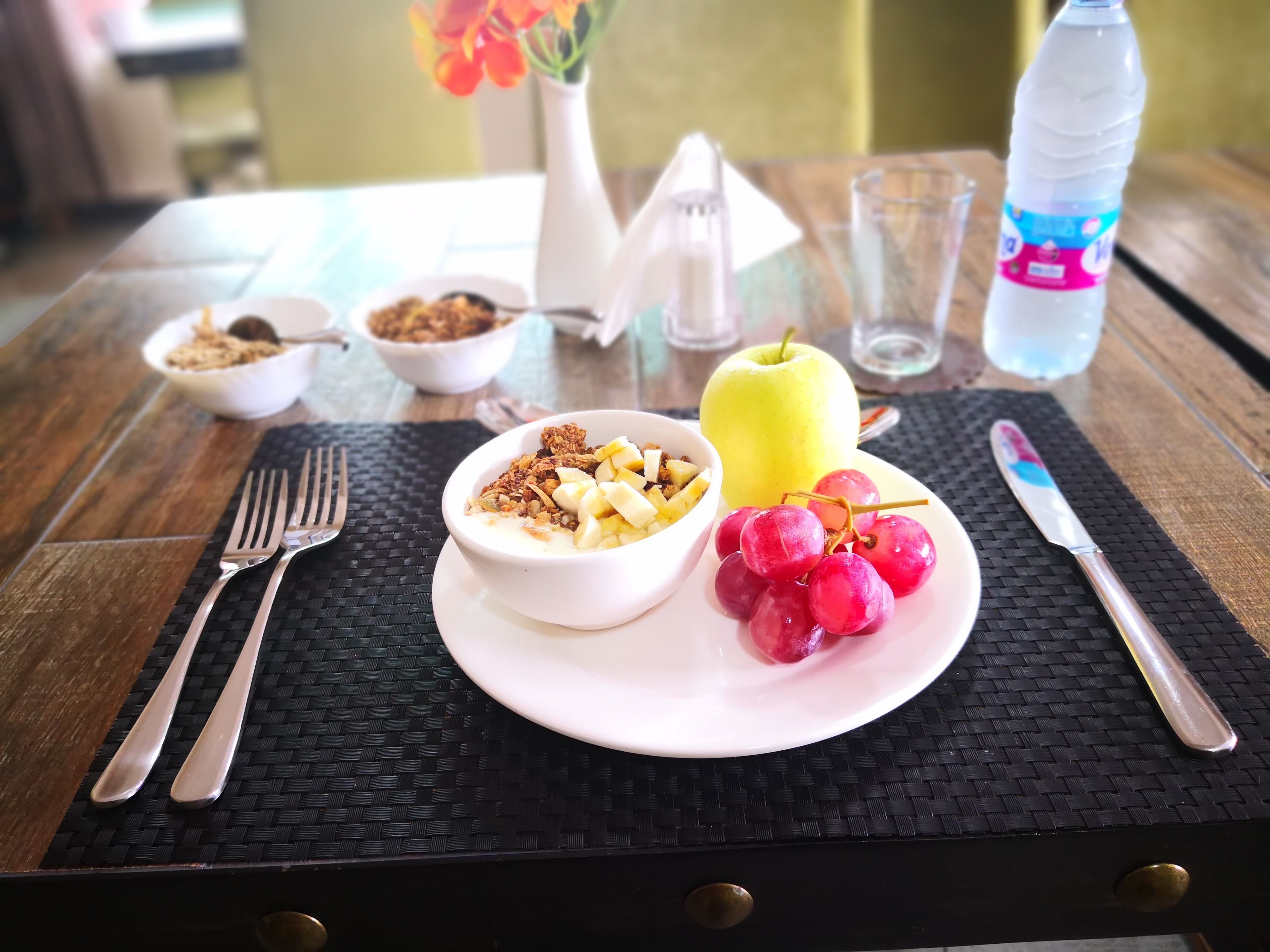 Desayuno con un tazón de cereales, un plato de uvas y una manzana, y una botella de agua sobre una mesa de madera.
