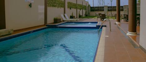 A swimming pool with clear water, surrounded by a tiled deck, lounge chairs, and a pergola structure.