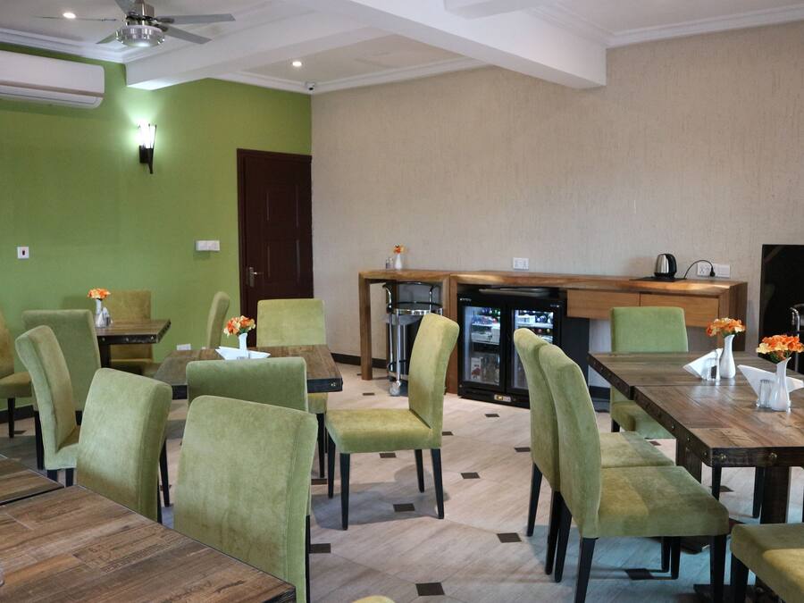 A hotel dining area with green walls, a wooden table, and matching chairs. There is a refrigerator and a coffee machine in the background.