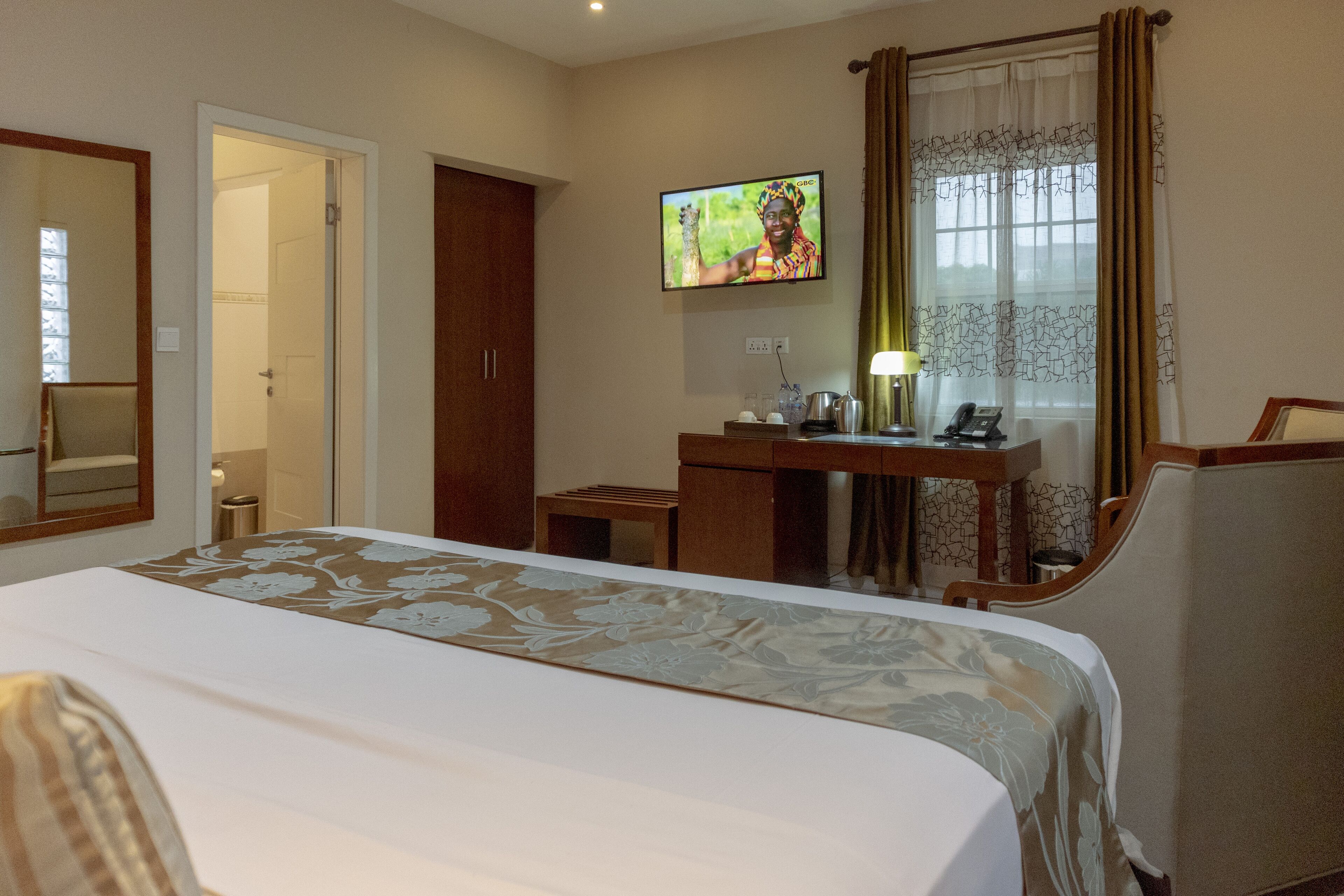 A hotel room with a large bed, a desk with a lamp, a television, and a window with curtains.