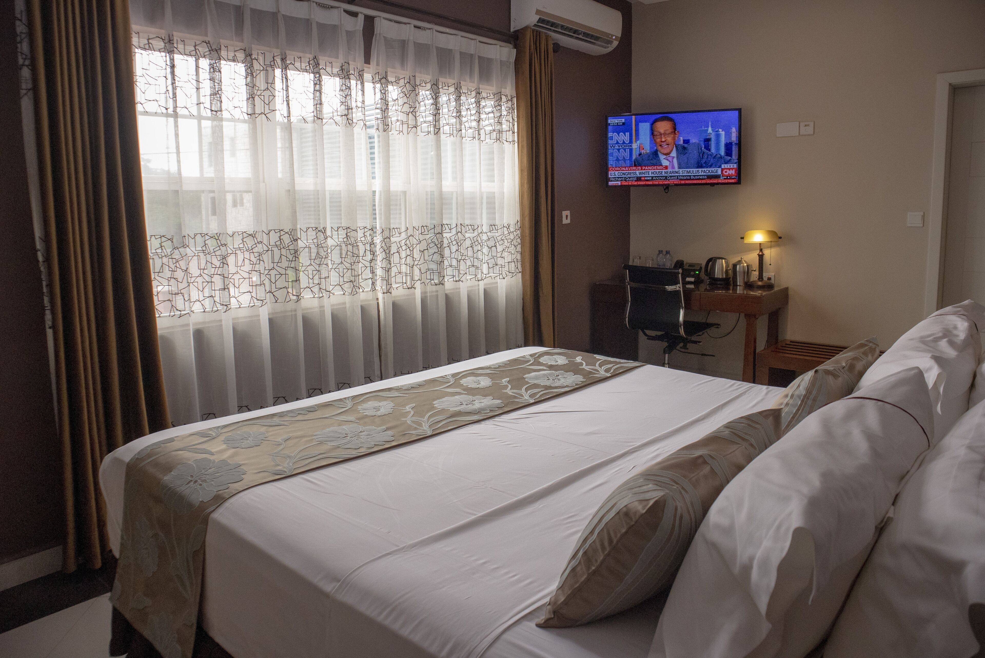 A hotel room with a bed, a desk with a computer, a TV on the wall, and a window with curtains.