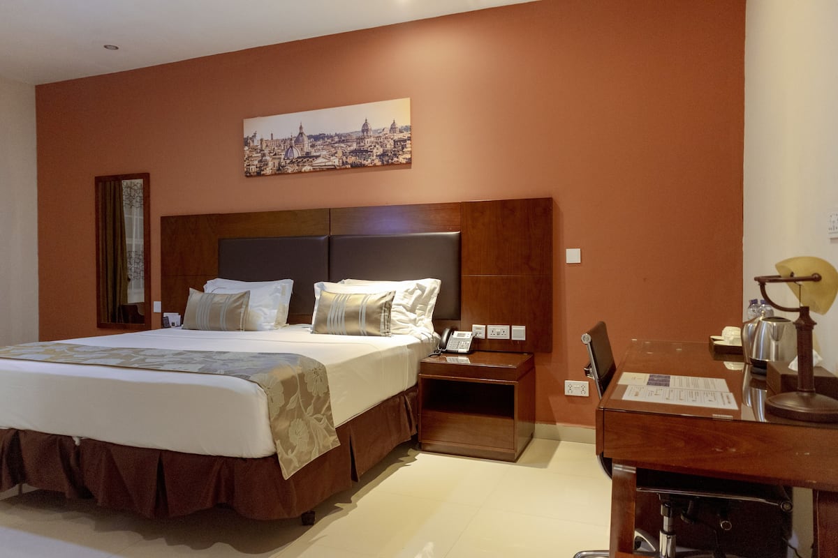 A hotel room with a large bed, a wooden nightstand, a desk with a lamp, and a cityscape painting on the wall.