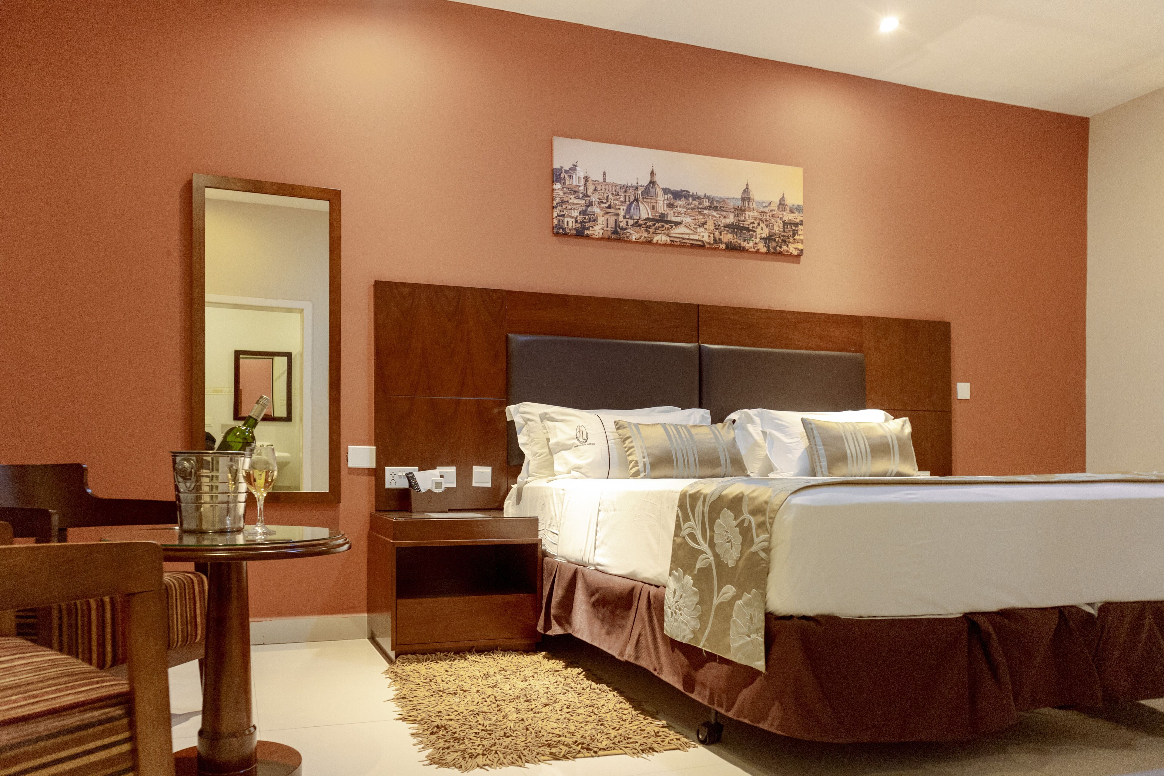 A hotel room with a bed, a wooden nightstand, a table with a bottle and glass, a chair, and a cityscape painting on the wall.