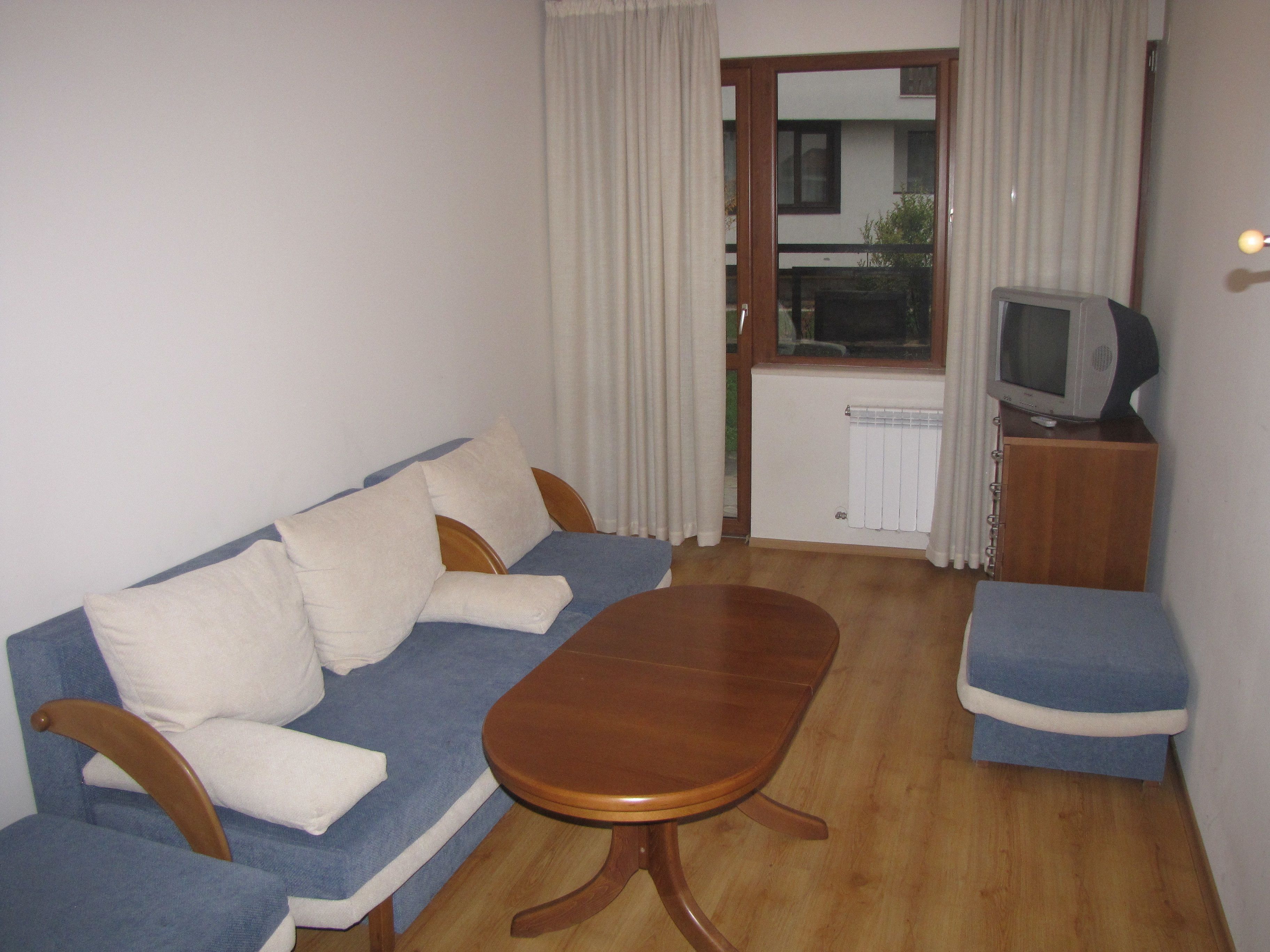 Un petit salon avec un canapé, une table basse en bois, un téléviseur posé sur un meuble en bois et une fenêtre avec des rideaux.