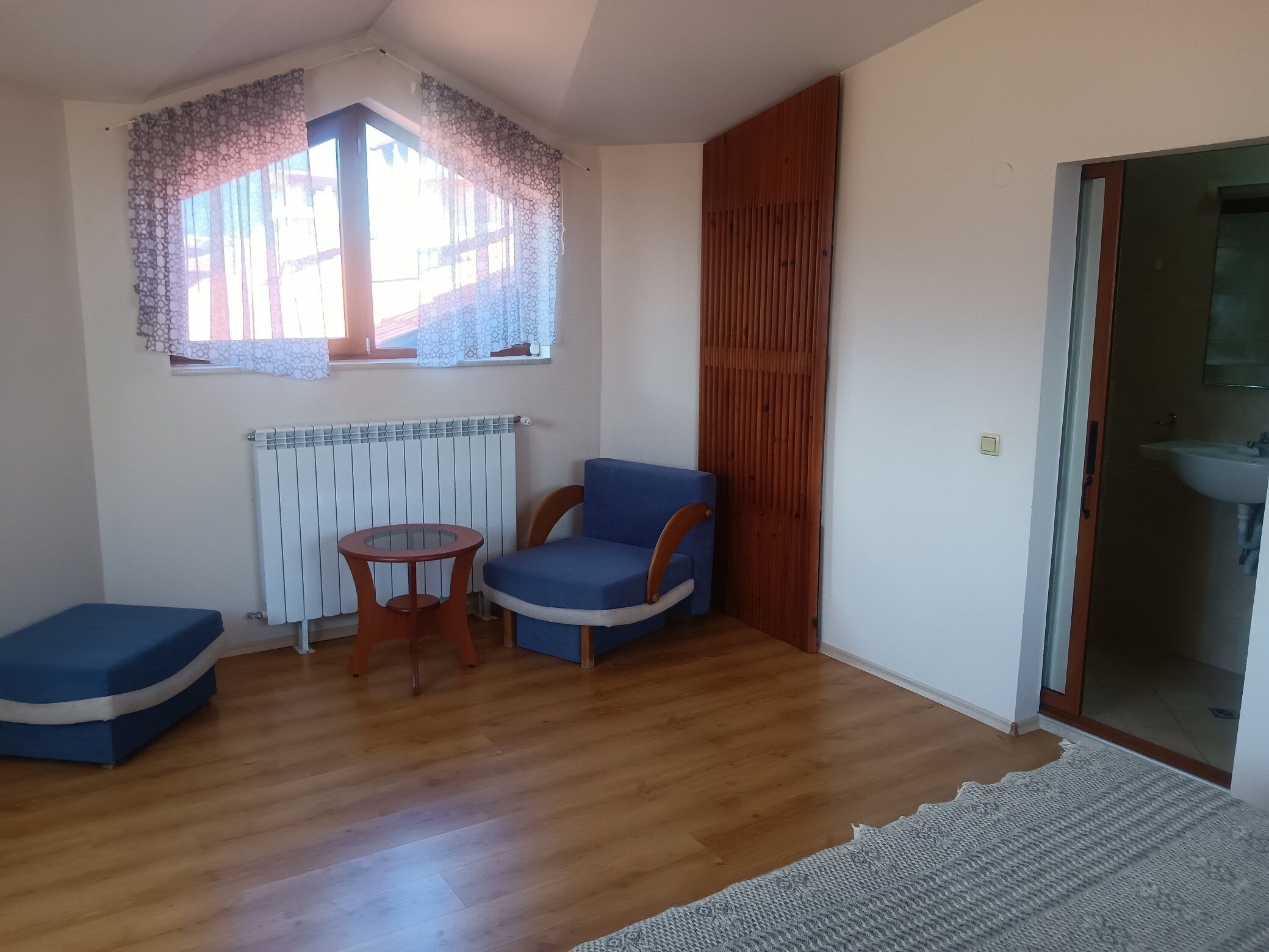 A small room with a wooden floor, a blue chair, a small table, a radiator, a window with patterned curtains, and a door leading to a bathroom.