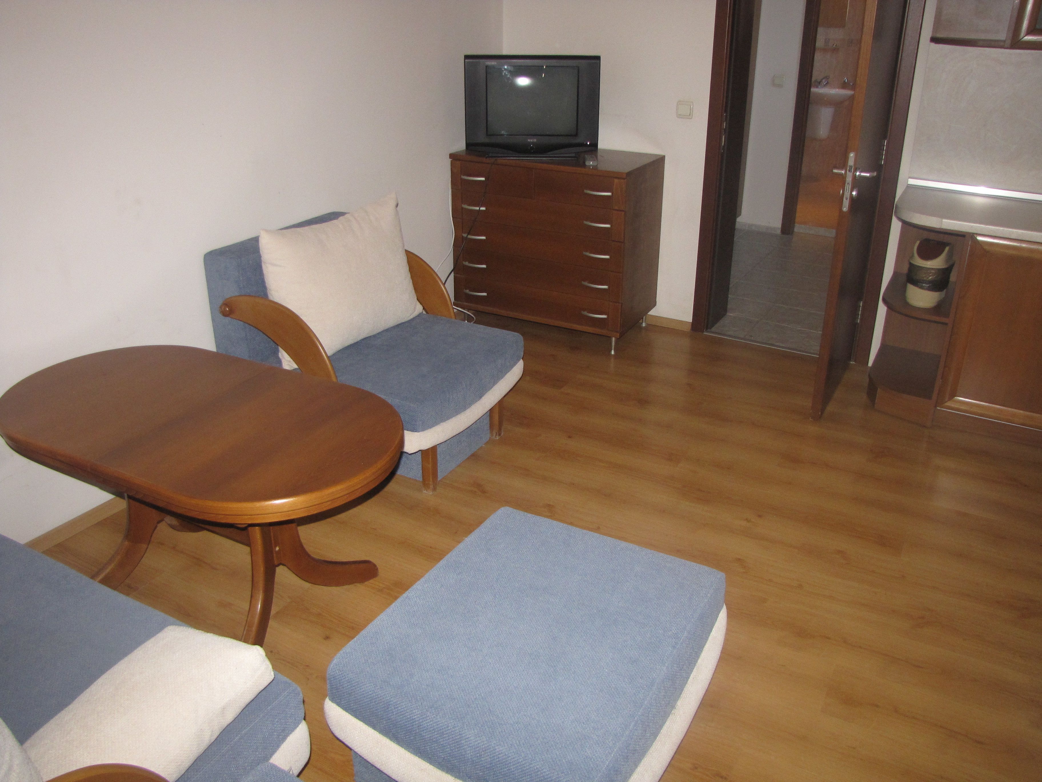 Un pequeño espacio de estar con una mesa de madera, dos sillones azules, un televisor sobre un mueble de madera y una zona de cocina con un fregadero y armarios.