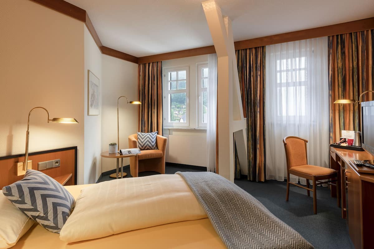 A hotel room with a bed, a desk, a chair, a lamp, and a window with curtains.