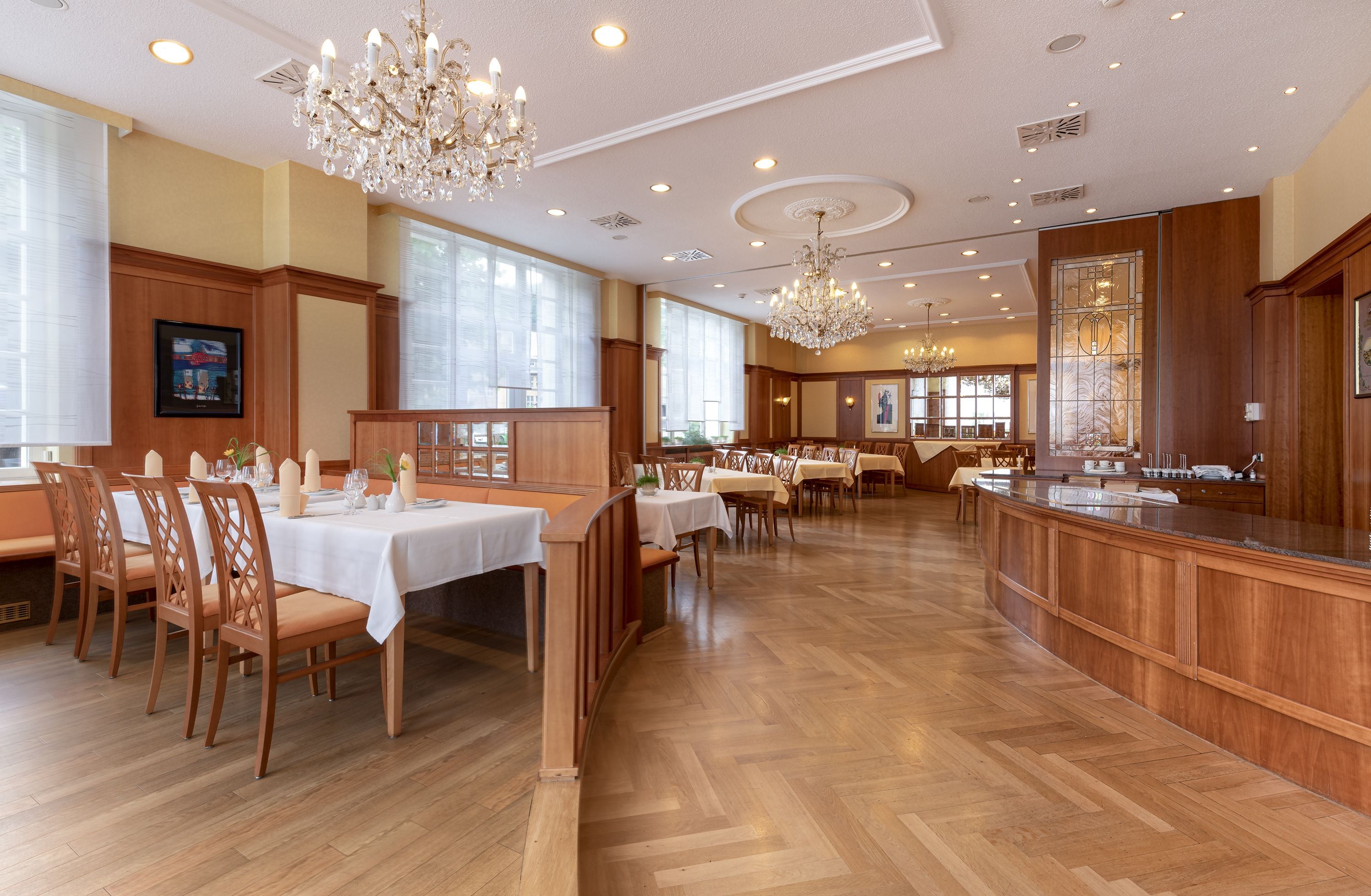 A spacious dining area with wooden flooring, white tablecloths, and elegant chandeliers.