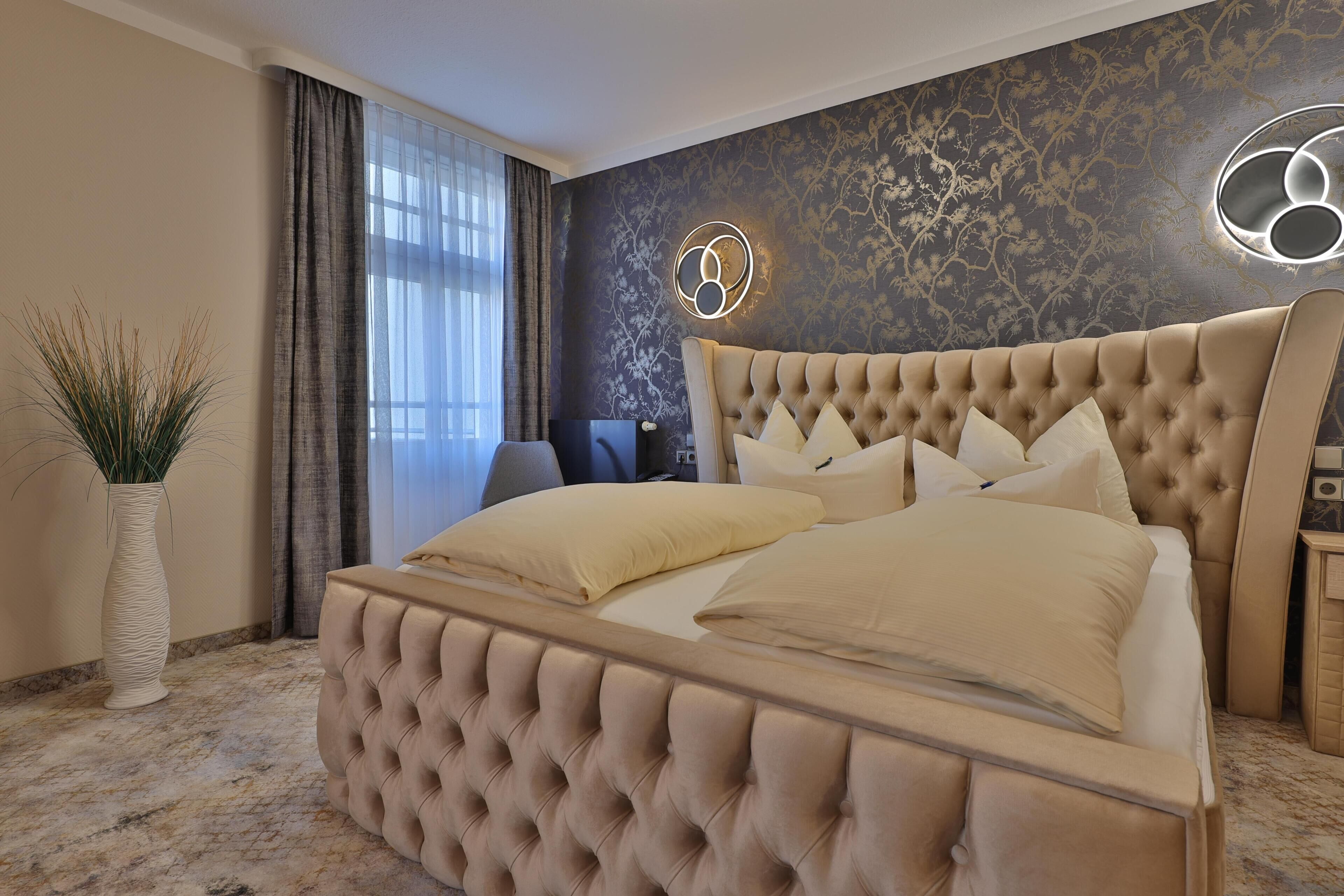Un dormitorio con cabecera capitonada, una cama grande con muchas almohadas, un florero con plantas y una lámpara fijada a la pared.