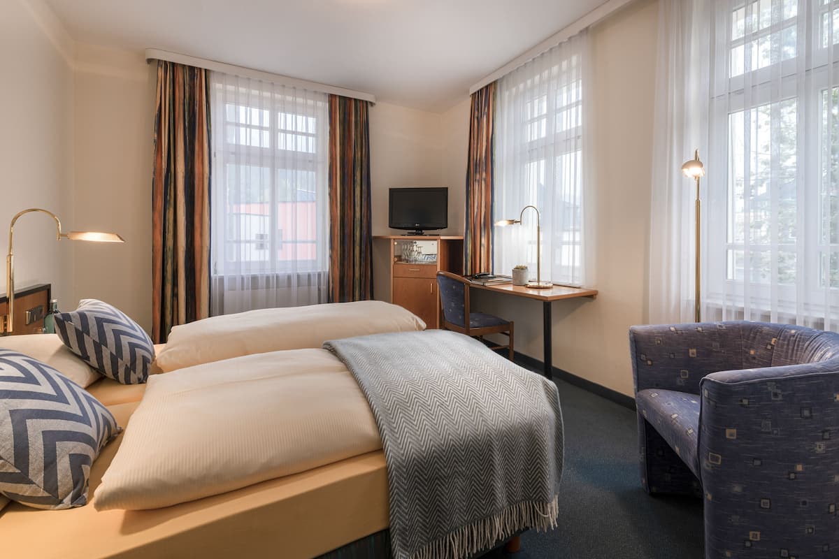 A hotel room with two beds, a desk, a chair, a television, and large windows with curtains.