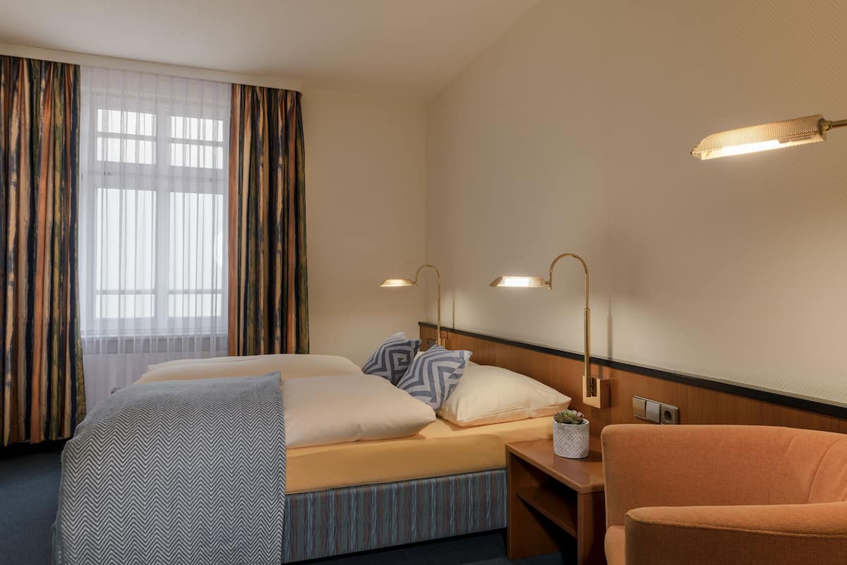 A hotel room with a bed, two bedside lamps, a chair, a small table, and a window with curtains.