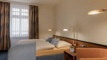 Ein Hotelzimmer mit einem Bett, zwei Nachttischlampen, einem Sessel, einem kleinen Tisch und einem Fenster mit Vorhängen.