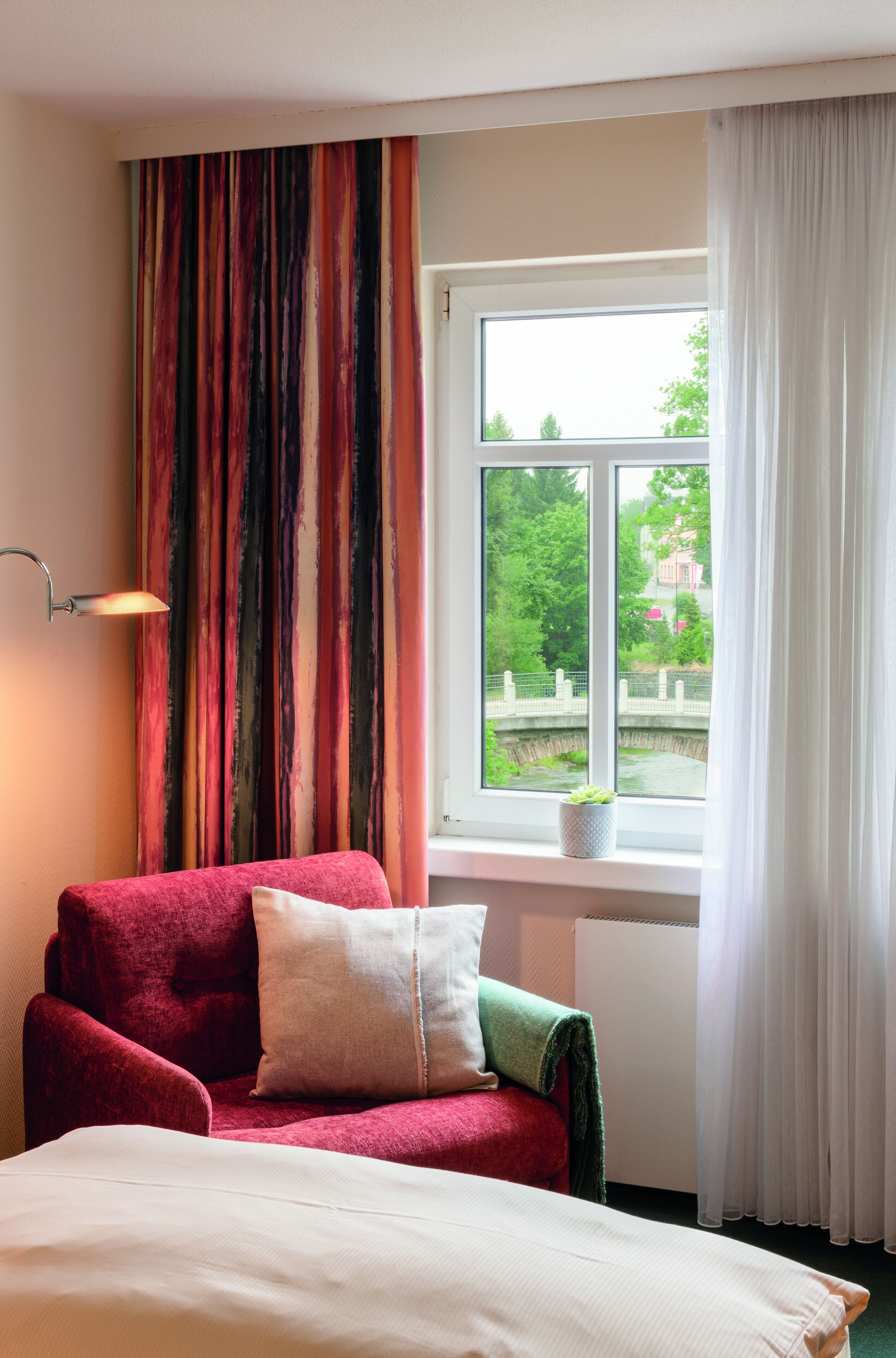 A room with a bed, a red armchair, a window with a view of greenery, and a lamp.