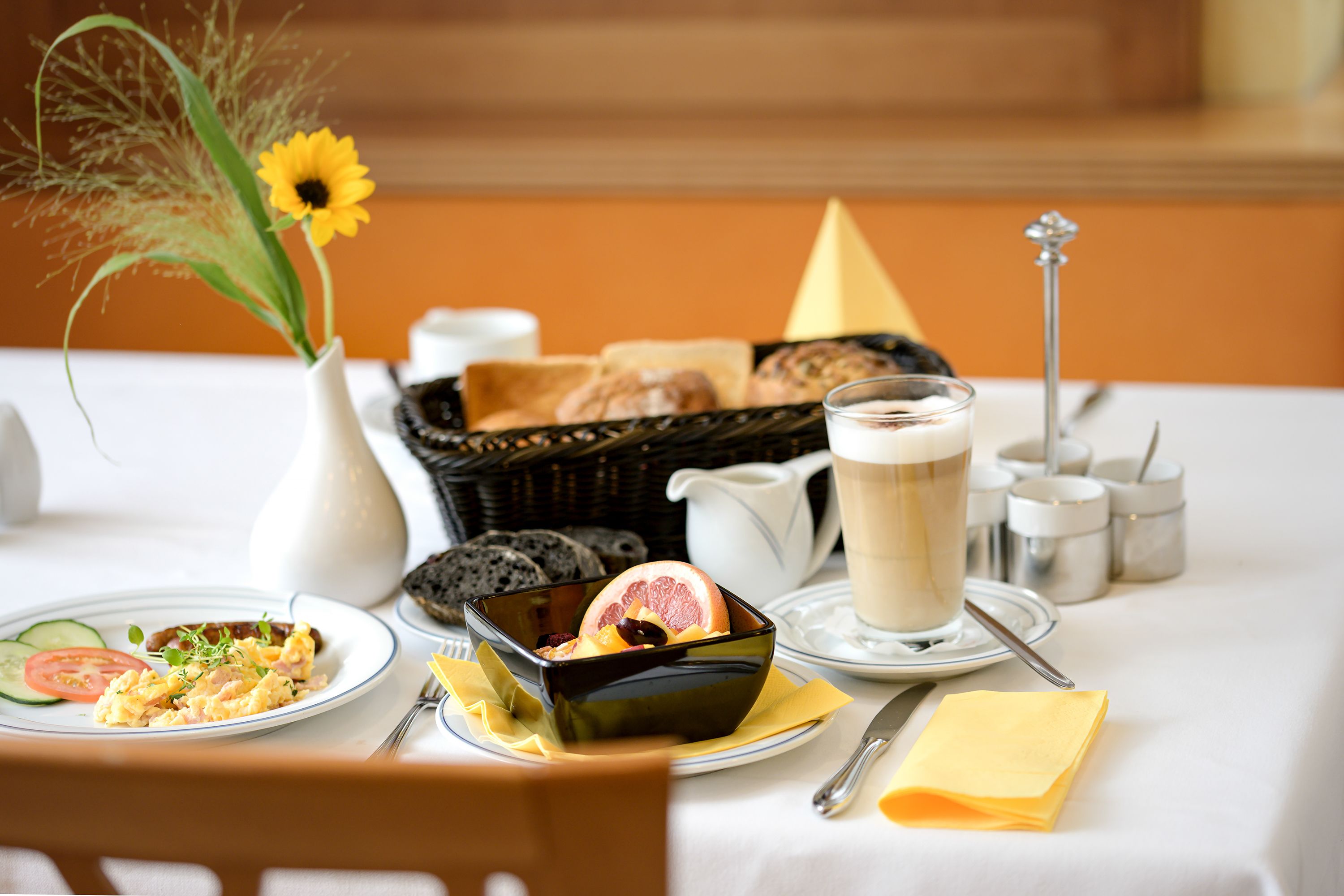 A breakfast setting with a plate of scrambled eggs, a bowl of fruit, a cup of coffee, and a vase with a sunflower.
