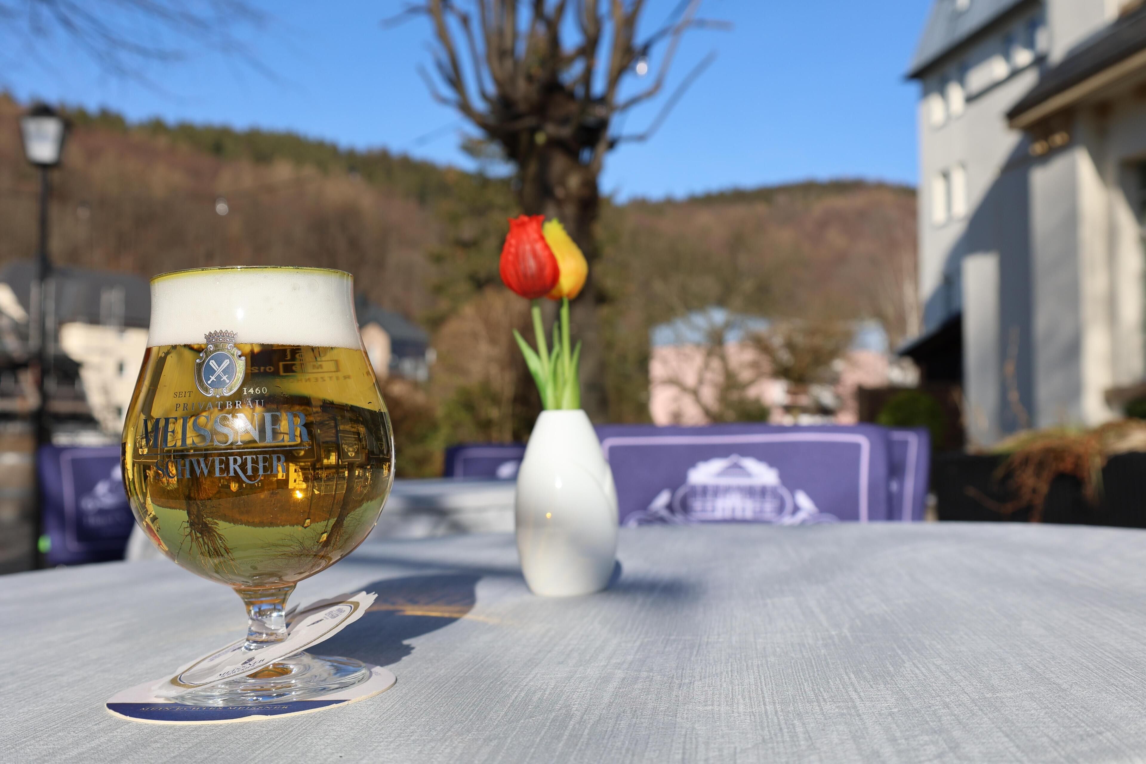 Um copo de cerveja sobre uma mesa, com uma tulipa e um vaso branco ao fundo.