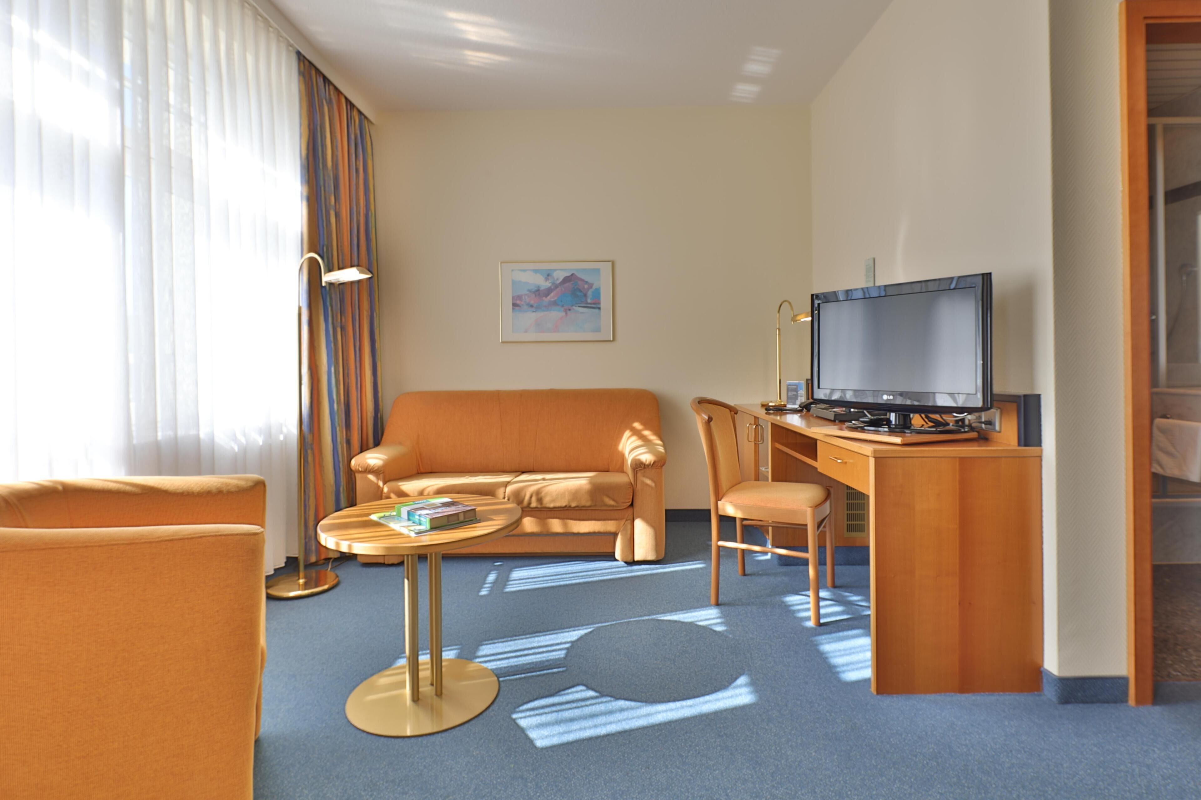 Habitación de hotel con sofá, escritorio con computadora, lámpara y un cuadro en la pared.